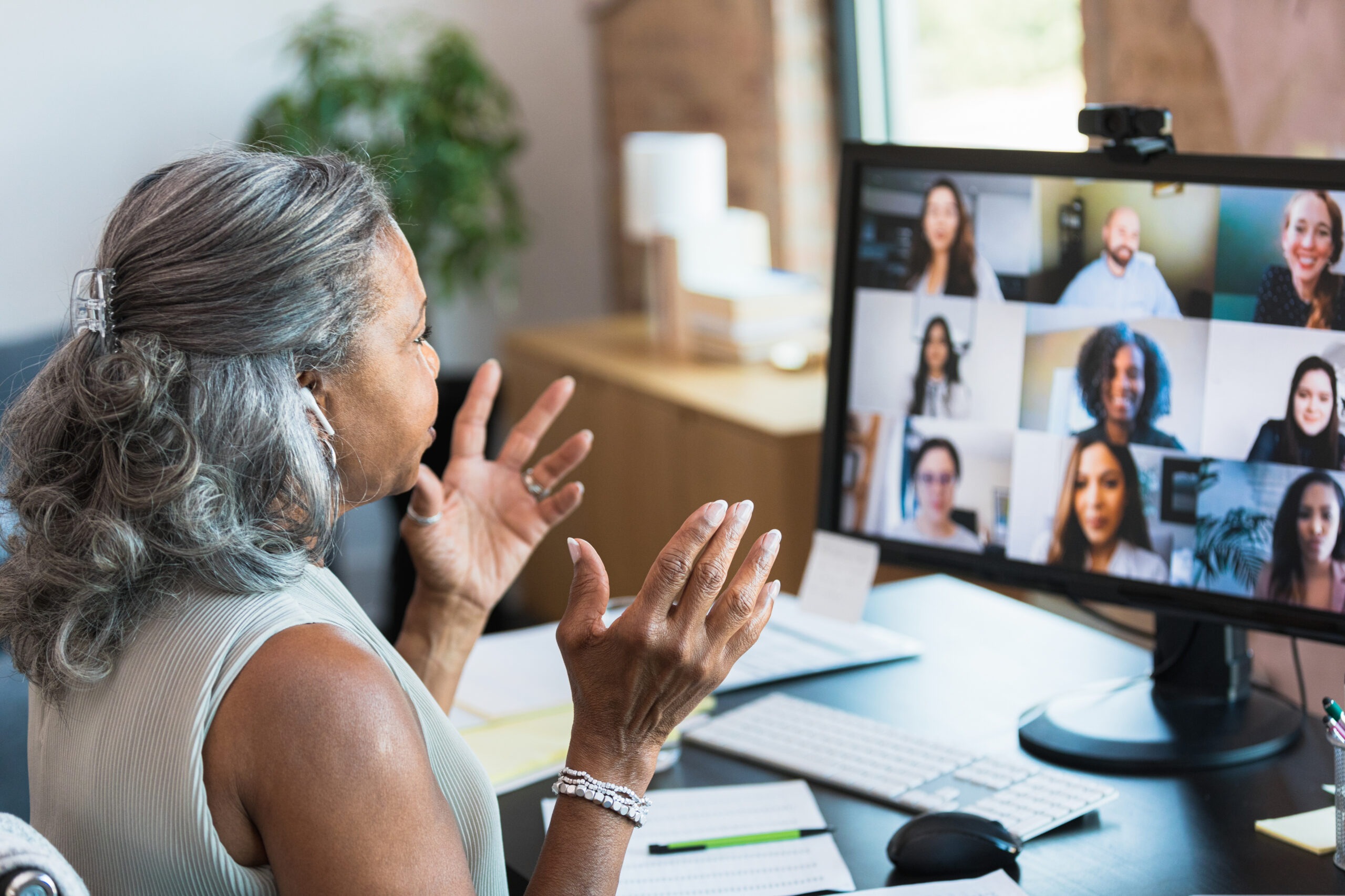 A senior businesswoman gestures as she facilitates a homeowners workshop while working from home. She is talking with the homebuyers workshop participants during a video conference.