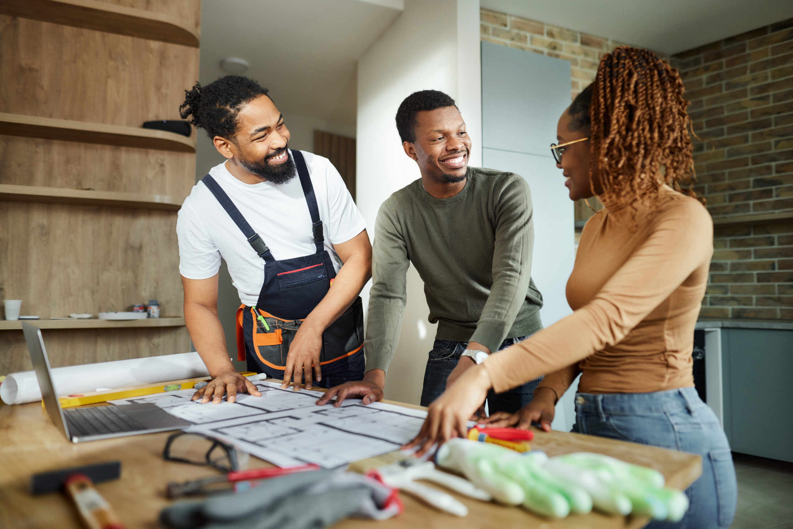 Happy African American couple talking while analyzing blueprints with manual worker in their home